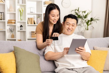 Online-Shopping, Paar sitzt auf Sofa mit Kreditkarte und digitalem Tablet im gemütlichen Wohnzimmer.