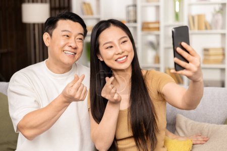 Foto de Feliz pareja asiática tomando selfie en casa. Hombre y mujer sonrientes posando para una foto móvil alegre en un ambiente elegante de la sala de estar, encarnando felicidad y estilo de vida moderno. - Imagen libre de derechos