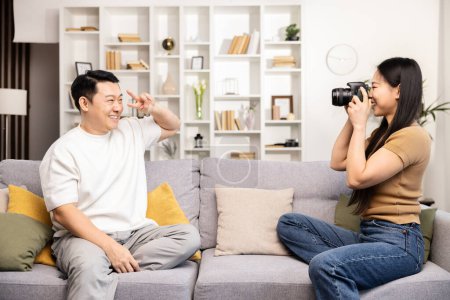 Téléchargez les photos : Un homme asiatique pose ludique tandis qu'une femme prend sa photo à l'intérieur d'un salon confortable, capturant un moment franc de joie et de passe-temps. - en image libre de droit