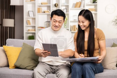 Téléchargez les photos : Couple assis ensemble sur un canapé, engagé dans la paperasserie et tablette numérique dans un salon confortable. - en image libre de droit