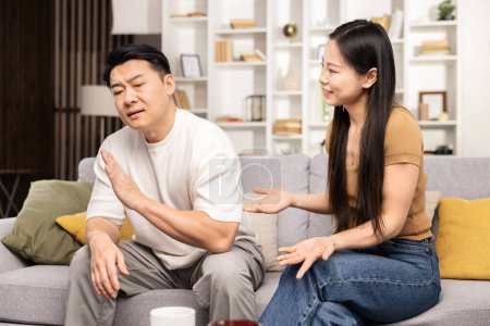 Reifer asiatischer Mann gestikuliert während des Streits, junge Asiatin erklärt, Paarstreit, Beziehungsprobleme, emotionaler Stress, Wohnzimmer im Haus, Konfliktlösung