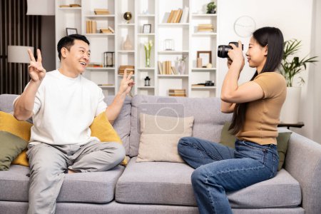 Foto de Un hombre alegre posa con un signo de paz mientras una mujer se centra en tomar su fotografía con una cámara réflex digital en una acogedora sala de estar. - Imagen libre de derechos