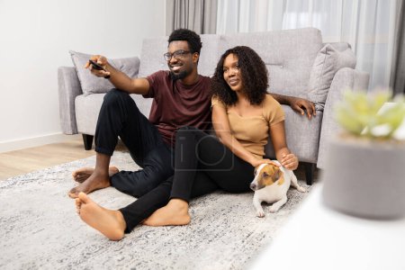 Téléchargez les photos : Couple heureux se détendre sur le sol avec chien regarder la télévision dans le salon moderne - en image libre de droit