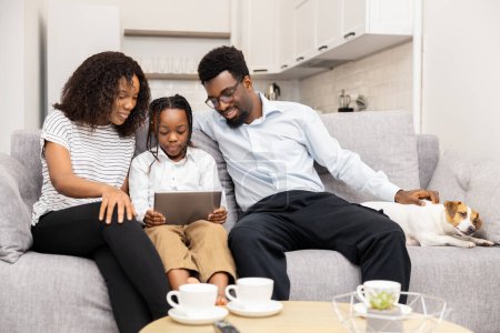 Téléchargez les photos : Famille afro-américaine heureuse utilisant la tablette sur le canapé avec chien de compagnie à la maison, convivialité détendue - en image libre de droit