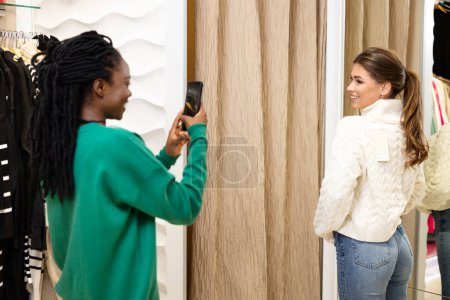 Téléchargez les photos : Joyeuse journée shopping : Jeune femme afro-américaine prend une photo de son ami essayant de nouveaux vêtements dans une boutique moderne - en image libre de droit