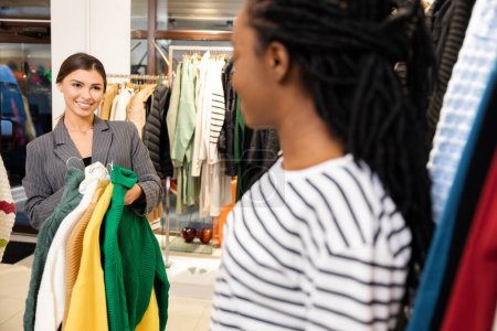Foto de Amistoso asistente de compras que ayuda al cliente en la tienda boutique, mujeres sonrientes que eligen ropa de moda, experiencia de venta al por menor agradable, moda y estilo - Imagen libre de derechos