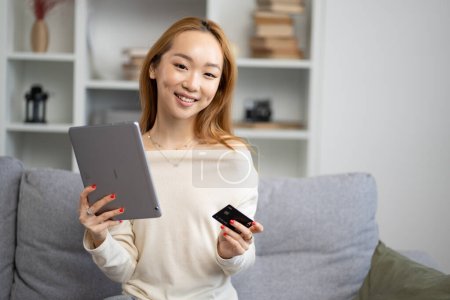 Junge Asiatin mit Tablet und Kreditkarte auf dem Sofa, Lächeln zu Hause, Online-Shopping, Einsatz moderner Technologie, komfortables Leben