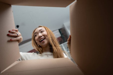Foto de Mujer feliz Unboxing: Joven asiática sonriendo mientras abre una caja de cartón, capturando emoción y alegría en los momentos cotidianos - Imagen libre de derechos