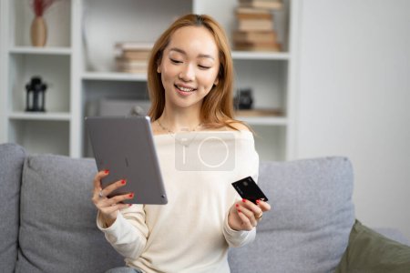 Jeune femme asiatique faisant du shopping en ligne sur tablette à la maison, souriant alors qu'elle détient une carte de crédit, confortable et confortable cadre de salon