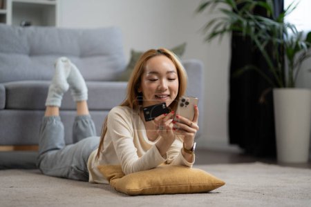Junge Frau genießt Online-Shopping auf dem Smartphone zu Hause, lächelnde asiatische Frau mit Kreditkarte, bequemes Liegen, moderne Wohnzimmereinstellung.