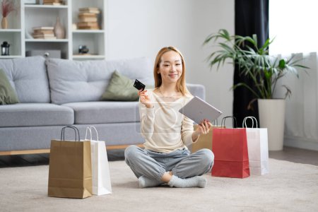 Junge Frau genießt Online-Shopping, sitzt mit Tablet, Kreditkarte und bunten Einkaufstaschen im eleganten Wohnzimmer auf dem Boden.
