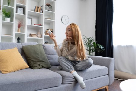 Téléchargez les photos : Jeune femme asiatique se relaxant sur le canapé à la maison, souriant et tenant une télécommande. Salon contemporain avec un décor élégant. Confort, Loisirs, Thème technologique. - en image libre de droit