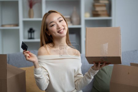 Glückliche Asiatin mit Kreditkarte und Box im Wohnzimmer, Online-Shopping, Konzept für den Umzugstag