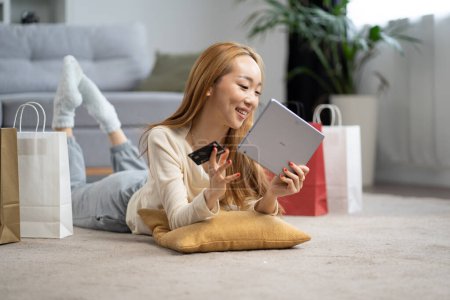 Téléchargez les photos : Joyeux jeune femme faisant du shopping en ligne à la maison, souriant alors qu'elle détient une carte de crédit et une tablette, sacs à proximité. Expérience e-commerce confortable. - en image libre de droit