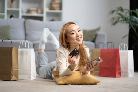 Happy Young Woman Shopping en ligne à la maison, tenant la carte de crédit et le téléphone portable avec des sacs à provisions autour. Concept de commerce électronique, Achats en ligne, Consommation.