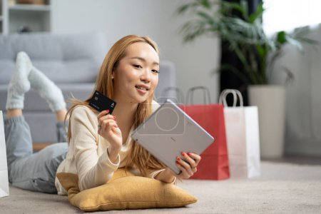Junge Asiatin kauft zu Hause online ein, lächelt mit Kreditkarte in der Hand, gemütliche Casual-Indoor-Szene, Technologie und Lifestyle-Konzept