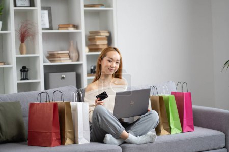 Happy Young Woman achats en ligne avec ordinateur portable et carte de crédit, entouré de sacs à provisions colorés sur un canapé confortable, mode de vie moderne et concept de commerce électronique