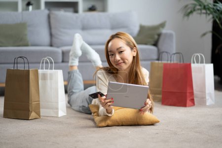 Foto de Joven mujer disfrutando de compras en línea en casa, sonriente señora asiática con tableta digital y tarjeta de crédito, rodeada de bolsas de compras en el piso de la sala de estar - Imagen libre de derechos