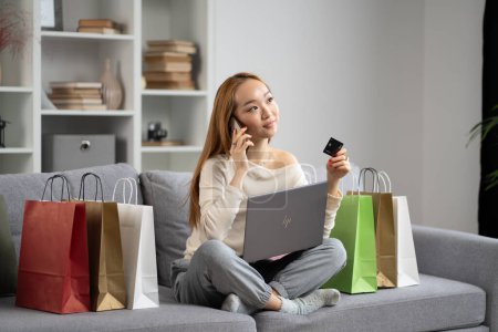 Jeune femme appréciant les achats en ligne et la conversation téléphonique, assis avec ordinateur portable et sacs à provisions colorés sur le canapé, heureux, détendu, Tech-Savvy, E-Commerce.