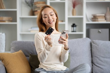 Photo for Happy Young Woman Using Smartphone And Holding Credit Card On Couch, Online Shopping, Mobile Banking In Modern Living Room. - Royalty Free Image