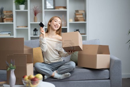 Foto de Mujer joven disfrutando de compras en línea, sosteniendo la tarjeta de crédito y sonriendo con cajas de cartón abiertas alrededor de ella en una acogedora sala de estar. - Imagen libre de derechos