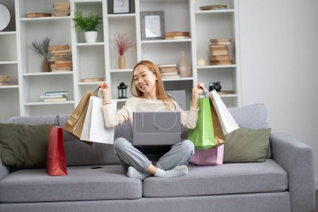 Happy Young Woman Shopping en ligne à la maison, sourire femme asiatique avec ordinateur portable et sacs à provisions colorés sur le canapé, concept de commerce électronique