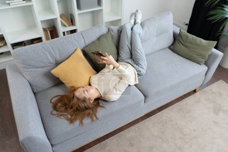 Mujer usando Smartphone y tarjeta de crédito en el sofá, Casual escena de estilo de vida en casa con elementos interiores modernos. Comodidad, ocio, uso de la tecnología en el hogar.