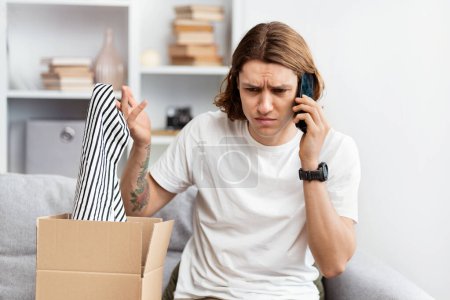 Young Man Unpacking Online Shopping Item, Looks Confused While Talking On Phone, Problem With Order, Suburban Home Background. Indoor, Daylight, Customer Service Issue, Consumer Rights.