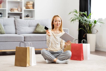 Junge Frau genießt Online-Shopping, sitzt mit Tablet, Kreditkarte und bunten Einkaufstaschen im eleganten Wohnzimmer auf dem Boden.