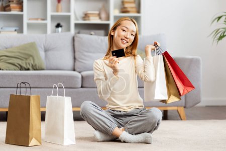 Téléchargez les photos : Joyeux jeune femme appréciant les achats en ligne à la maison, souriant femelle tenant carte de crédit et sacs à provisions colorés, concept de commerce électronique - en image libre de droit