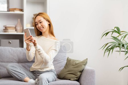 Téléchargez les photos : Femme asiatique heureuse utilisant un smartphone sur un canapé gris, style de vie décontracté, salon moderne, créateur de contenu, technologie à la maison - en image libre de droit