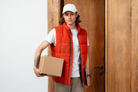 Confident Young Delivery Man In Red Vest Holding Package, Indoor Doorway Background. Professional, Reliable Delivery Service Concept.