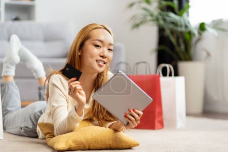 Junge Asiatin kauft zu Hause online ein, lächelt mit Kreditkarte in der Hand, gemütliche Casual-Indoor-Szene, Technologie und Lifestyle-Konzept
