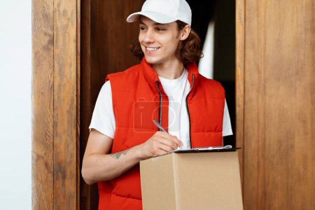 Photo for Friendly Delivery Man With Package Standing At Door, Smiling Service Worker In Red Vest And White Cap. Express Parcel Delivery, E-Commerce, Customer Satisfaction - Royalty Free Image