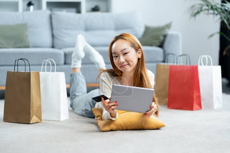 Jeune femme appréciant les achats en ligne à la maison, sourire dame asiatique avec tablette numérique et carte de crédit, entouré de sacs à provisions sur le plancher du salon