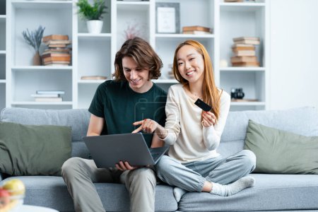 Glückliches Paar kauft zu Hause online ein. Junge Männer und Frauen sitzen lächelnd auf einer Couch, während sie in einem gemütlichen Wohnzimmer mit Kreditkarte und Laptop einkaufen.