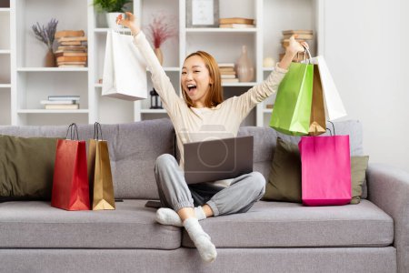 Joyeuse jeune femme célèbre le succès du shopping en ligne avec des sacs colorés, exprimant l'excitation et le bonheur dans le salon confortable.