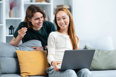 Photo for Happy Couple Shopping Online Together At Home. A Young Man Holding A Credit Card, Smiling Woman Browsing Laptop. Cozy, Connected, Leisure, E-Commerce Concepts - Royalty Free Image