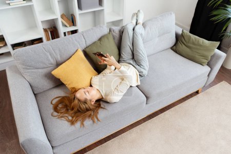 Mujer usando Smartphone y tarjeta de crédito en el sofá, Casual escena de estilo de vida en casa con elementos interiores modernos. Comodidad, ocio, uso de la tecnología en el hogar.
