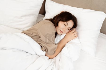 Erholsamer Schlaf: Eine junge Frau, die es sich in einem gemütlichen weißen Bett bequem macht und ein Gefühl der Entspannung und Ruhe ausstrahlt, perfekt für Themen über Wohlbefinden und gesunden Lebensstil.