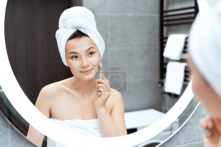 Junge Frau genießt Hautpflege-Routine im modernen Badezimmer, lächelt Spiegel mit Handtuch auf dem Kopf an und benutzt Gesichtsroller. Frisches, sauberes, Schönheitskonzept.