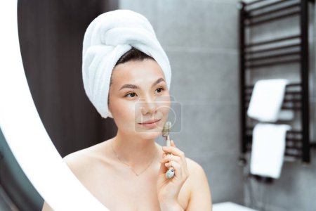 Junge Frau mit Jade-Roller im modernen Badezimmer: Eine gelassene Asiatin mit einem Handtuch auf dem Kopf benutzt eine Jade-Gesichtsrolle, die in einem stilvollen Badezimmer Ruhe und Selbstversorgung ausstrahlt.