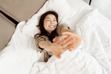Fröhliche Frau, die sich im Bett dehnt, morgens aufwacht, zu Hause glücklich ist. Lächelnde Weibchen genießen frischen Start in den Tag im hellen Schlafzimmer.