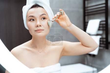 Femme utilisant le rouleau de jade dans la salle de bain moderne : Une image sereine et axée sur l'auto-soin mettant en vedette une femme asiatique avec une serviette sur la tête à l'aide d'un rouleau de jade sur son visage dans une salle de bain élégante.
