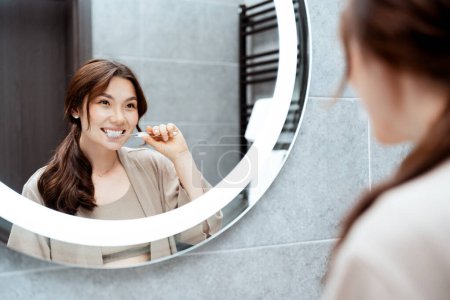 Foto de Mujer joven feliz cepillando los dientes en el baño moderno, reflejándose en el espejo con un interior elegante. Cuidado dental, rutina matutina y concepto de estilo de vida saludable. - Imagen libre de derechos