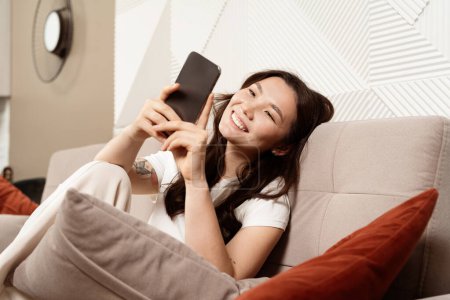 Téléchargez les photos : Jeune femme bénéficiant des médias sociaux sur Smartphone à la maison, allongé confortablement sur le canapé avec expression heureuse, intérieur moderne du salon - en image libre de droit