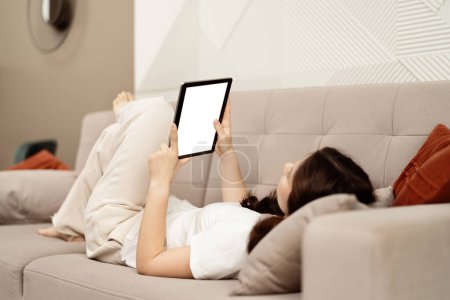 Foto de Mujer relajada usando la tableta en el sofá en casa. Una acogedora escena que representa a una joven tumbada en un sofá, navegando en una tableta digital en una elegante y moderna sala de estar. - Imagen libre de derechos