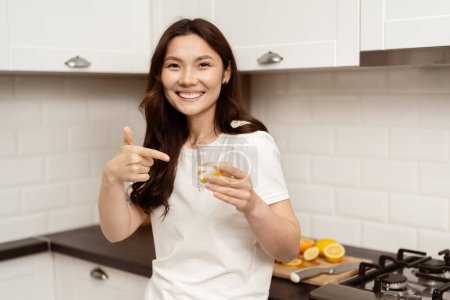 Téléchargez les photos : Jeune femme asiatique appréciant le verre d'eau dans la cuisine moderne, souriant heureusement, pointant du doigt sur le verre, mode de vie sain, bien-être, vie familiale décontractée - en image libre de droit