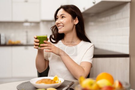 Foto de Young Woman Enjoying Healthy Breakfast, Smiling And Holding A Green Smoothie In A Modern Kitchen, Wellness And Healthy Lifestyle Concept - Imagen libre de derechos