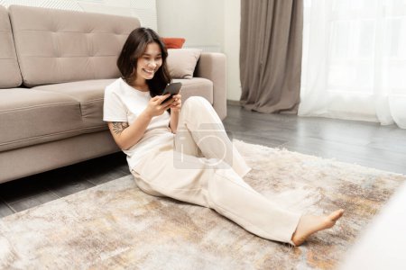 Foto de Mujer asiática joven que se relaja en casa, usando Smartphone en el piso de la sala de estar. Acogedor Interior Moderno, Tiempo Libre, Conectividad Digital. - Imagen libre de derechos